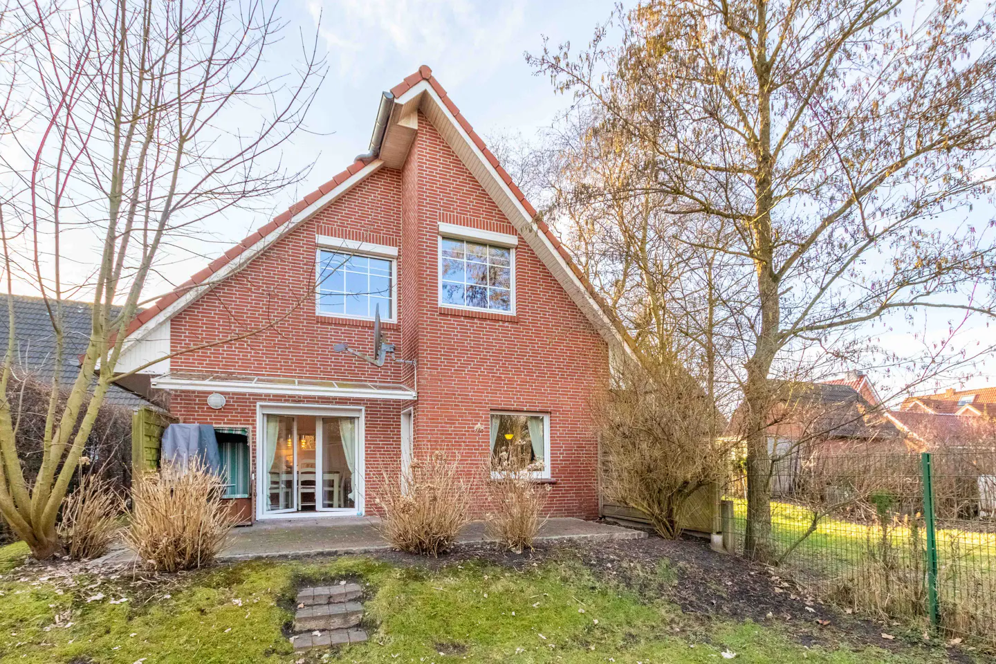 Backyard view of a two-story red brick house with a red roof, windows, and a patio with a glass door.