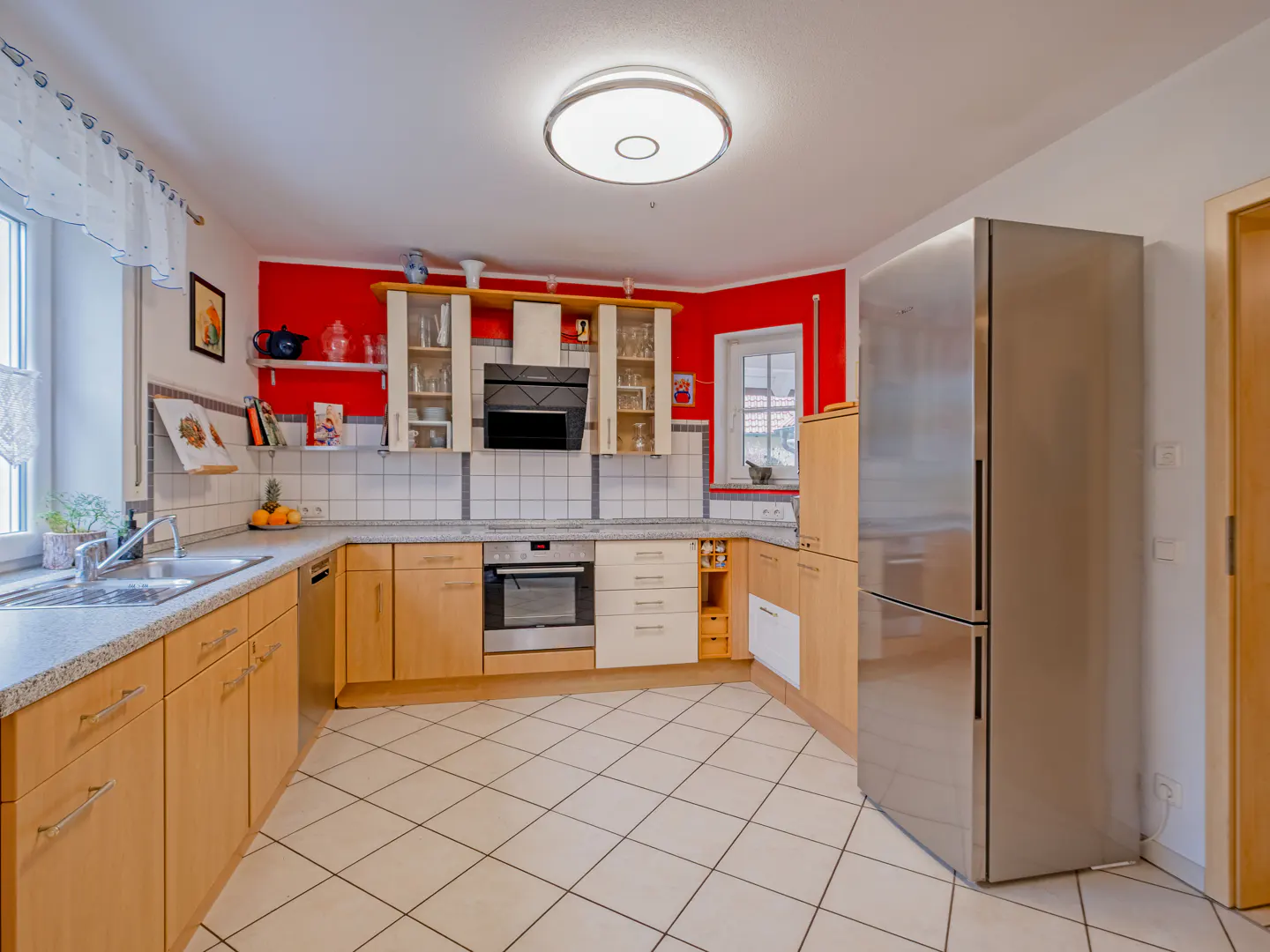 A bright kitchen with light wood cabinets, stainless steel refrigerator, and red accent wall. White tile floor and a window with a white curtain.