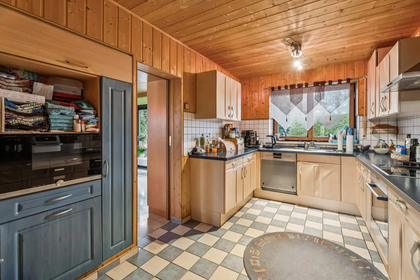 A kitchen with wood-paneled walls and ceiling, light wood cabinets, and a blue and white checkered floor.