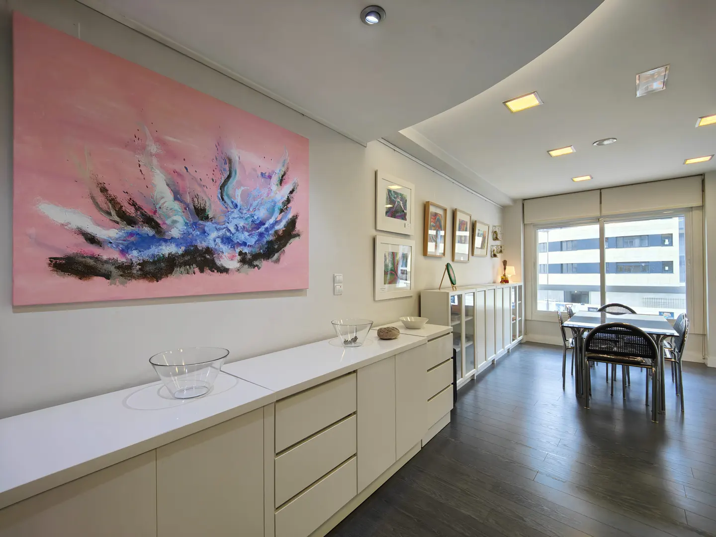 Bright living room with a pink abstract painting, white cabinets, and a dining table near a large window.
