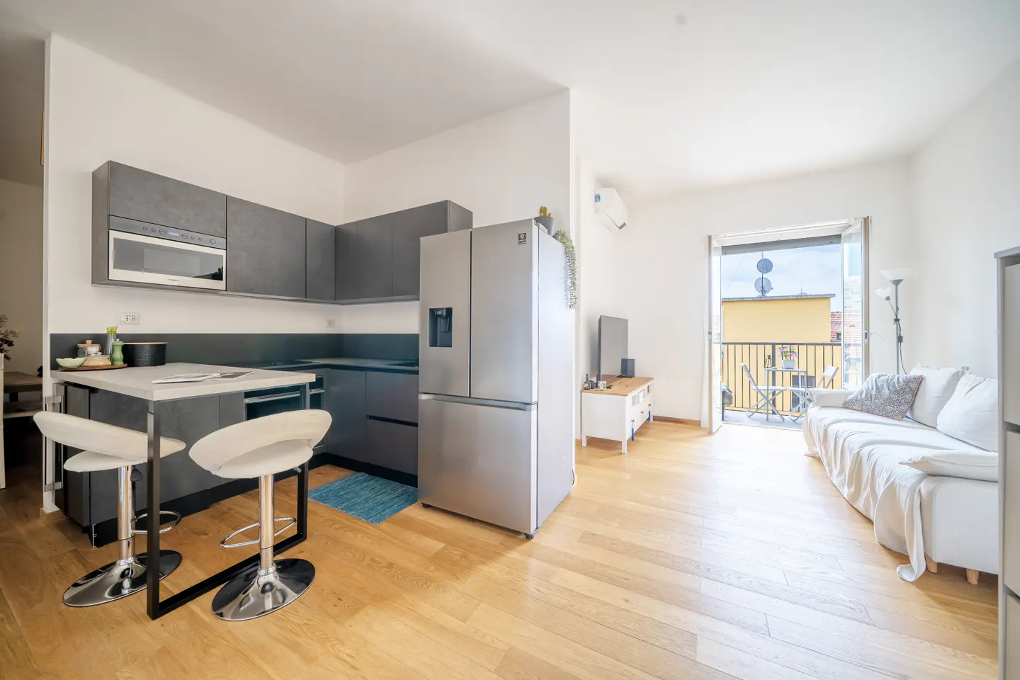 A bright, modern apartment interior with gray kitchen cabinets, a stainless steel refrigerator, and a living area with a white sofa and balcony.