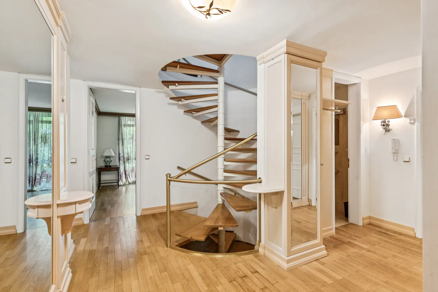 Bright foyer with a wooden spiral staircase, light wood floors, and white walls. Mirrored hall trees flank doorways.