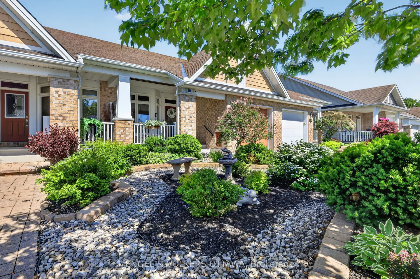 A tan brick bungalow with a red front door, stone landscaping, a small birdbath, and lush green bushes under a blue sky.