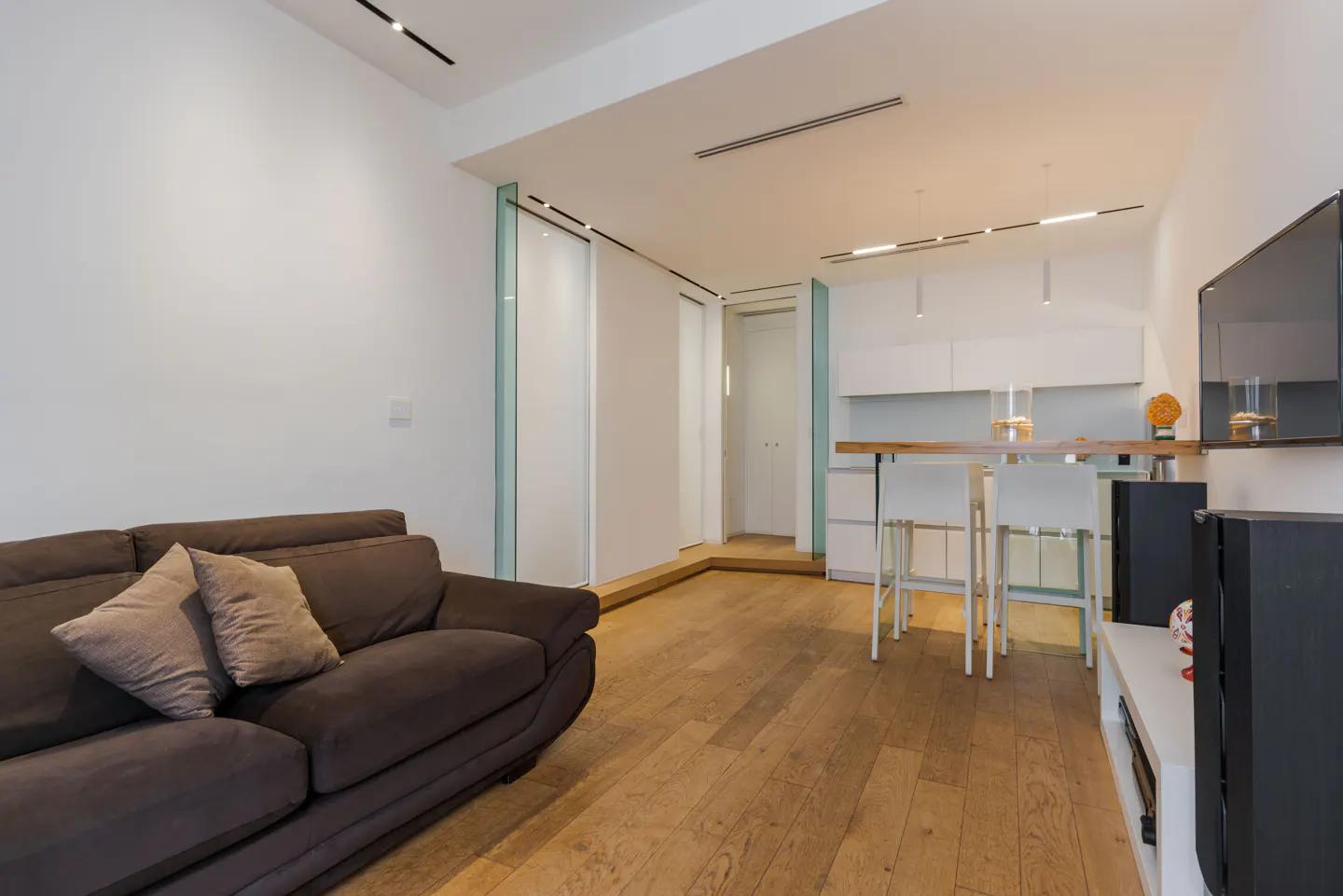 Living room with a brown sofa, wood floors, and a kitchen area with white cabinets and a breakfast bar.