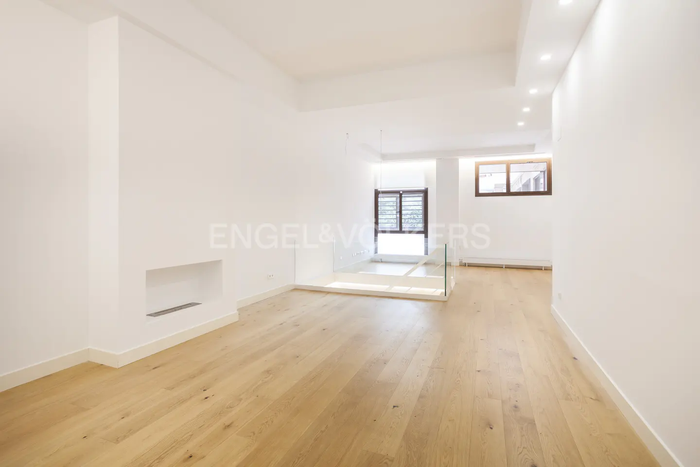 Bright, modern living room with light wood floors, white walls, and recessed lighting. A glass railing separates the space. Windows provide natural light.