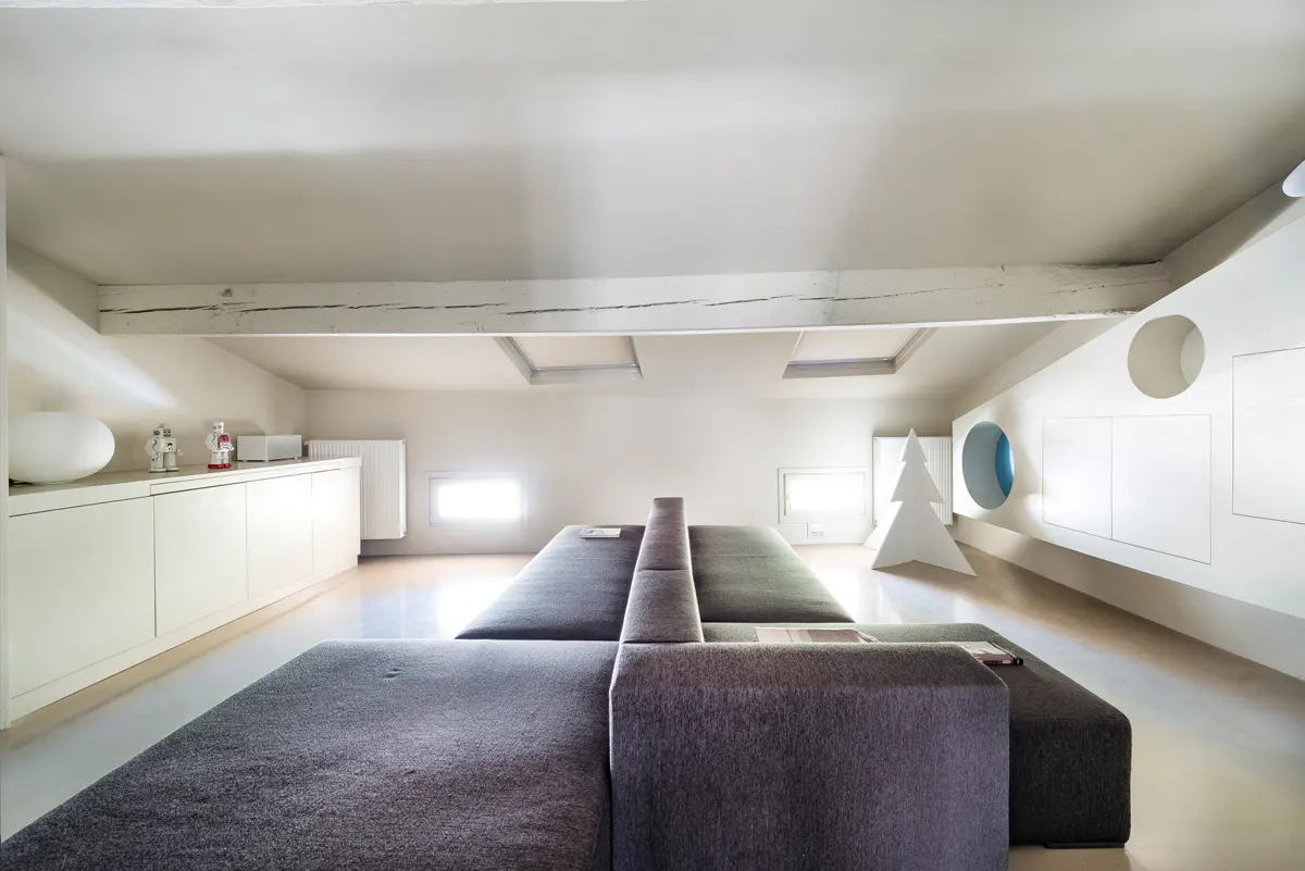 Attic living room with gray modular sofa, white walls, and a wooden beam. There are white cabinets, a radiator, and a white Christmas tree decoration.