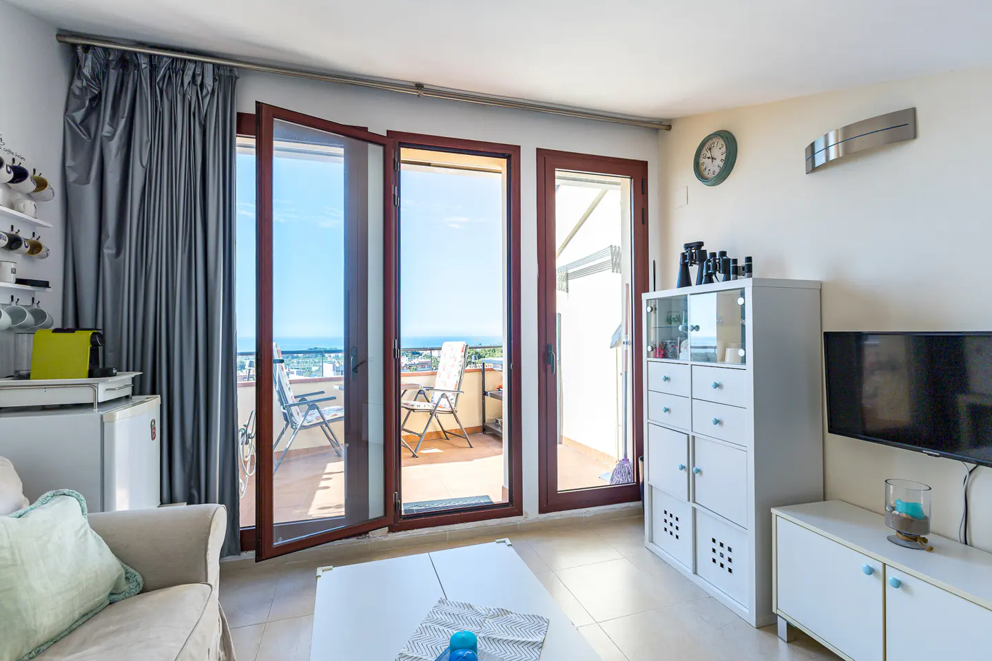 Bright living room with open balcony doors. Gray curtains, white furniture, and a view of the ocean.