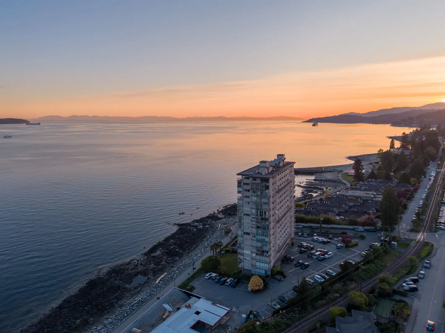 Aerial view of a tall building next to a rocky beach and ocean at sunset. Parking lot and street with cars visible.