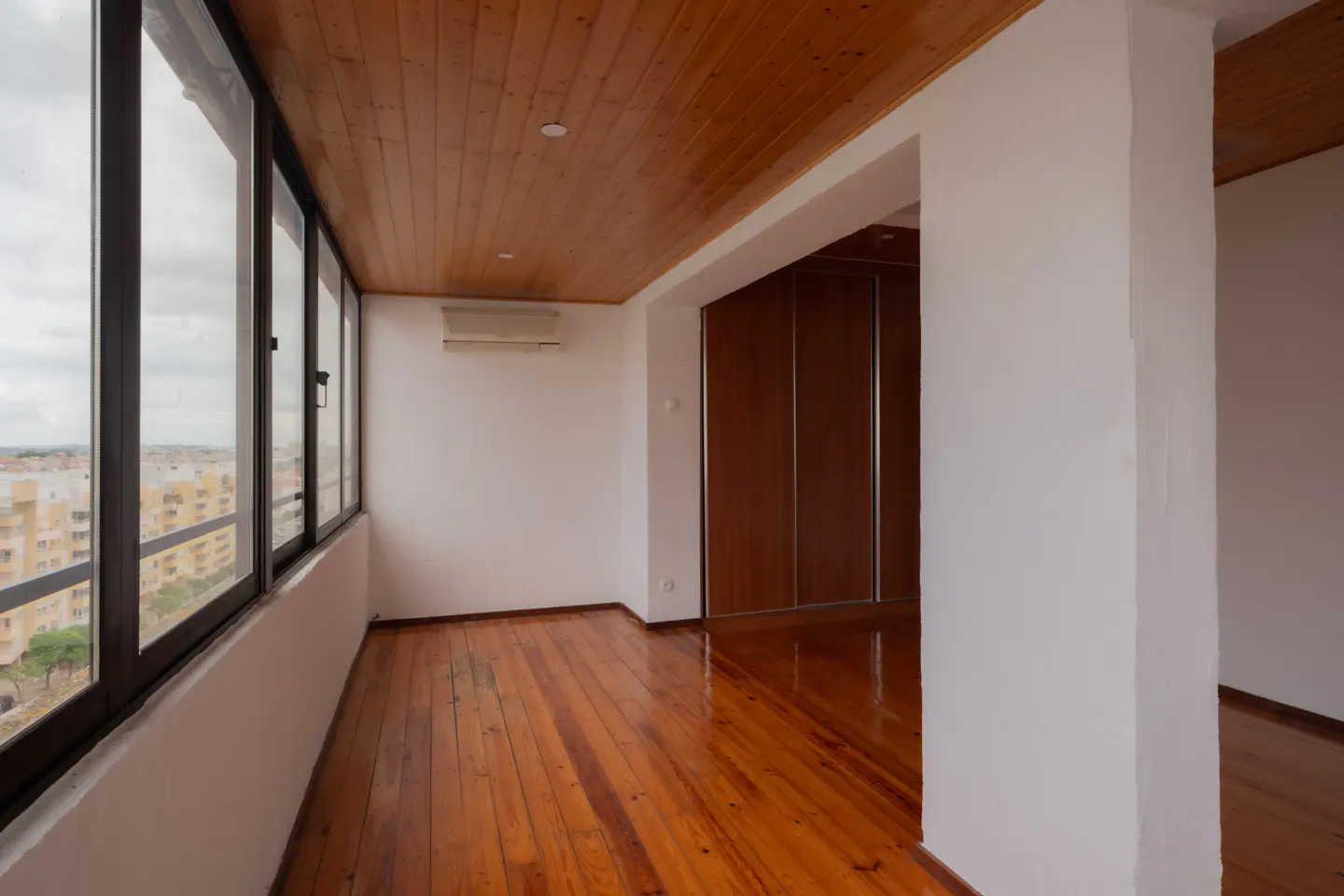Bright room with wood floors, ceiling, and large windows. White walls, and a view of a city.