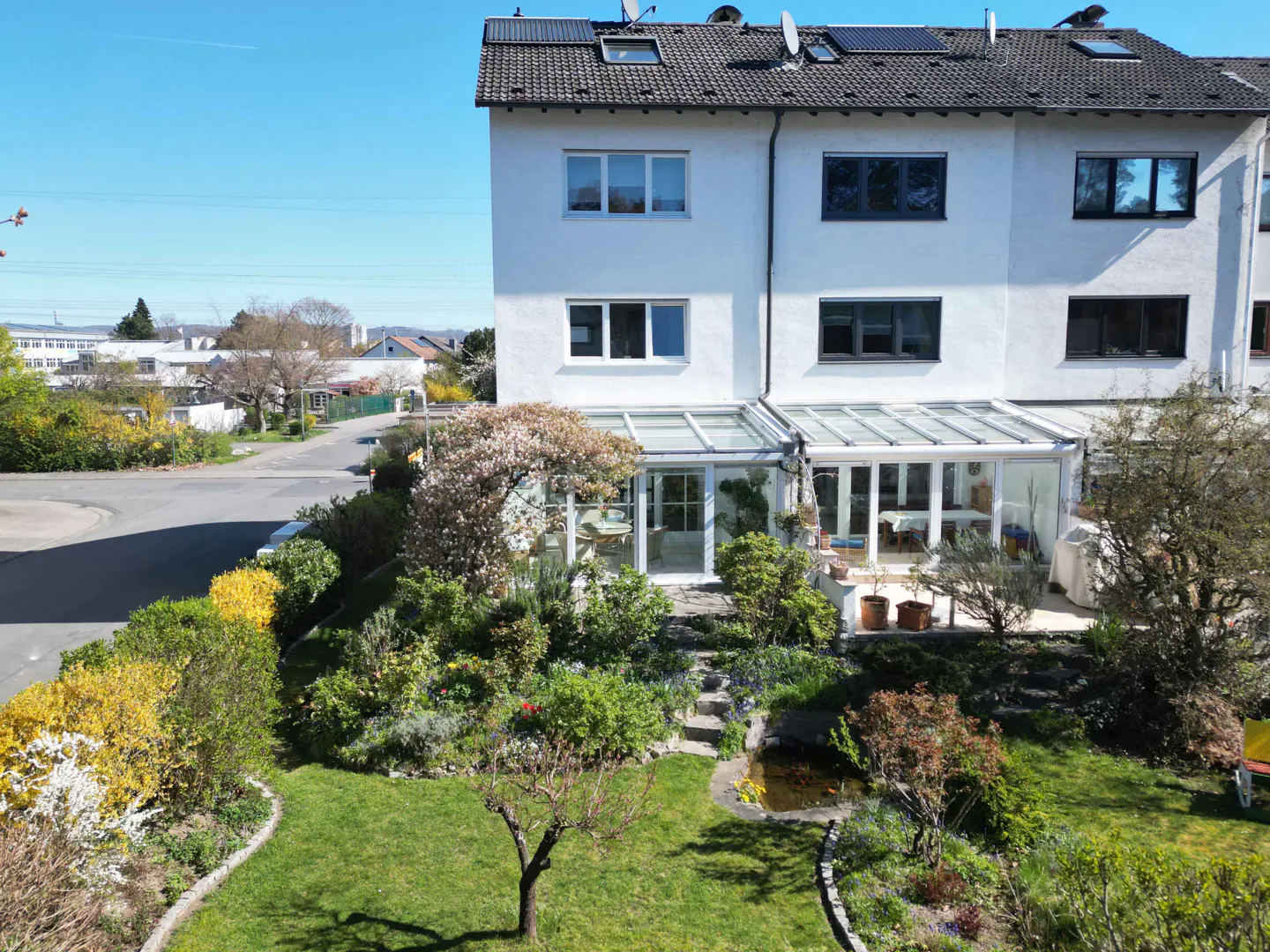 Two-story white townhouse with a glass-enclosed sunroom and a lush green garden with a small pond.
