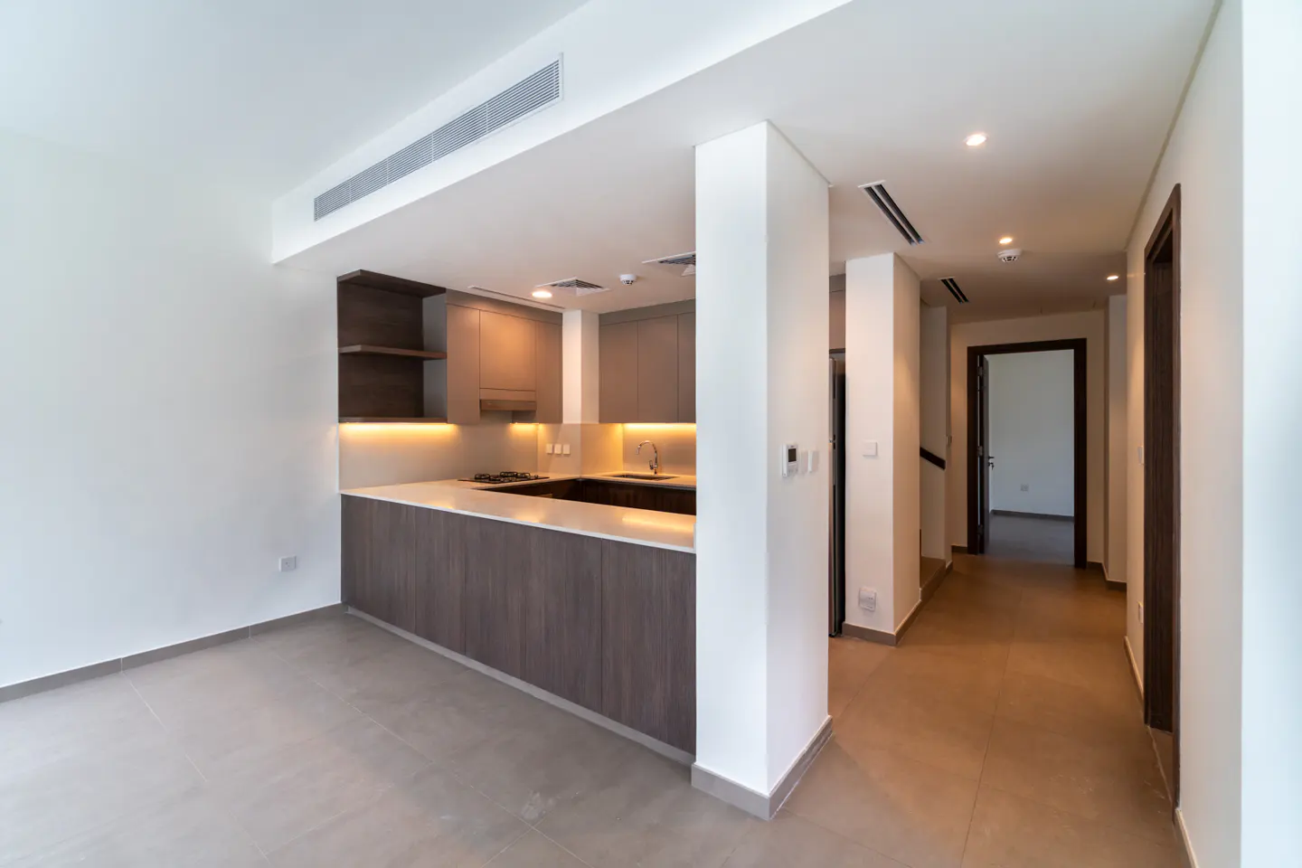 A bright, modern kitchen with gray cabinets, white countertops, and stainless steel appliances, next to a hallway with dark wood doors.