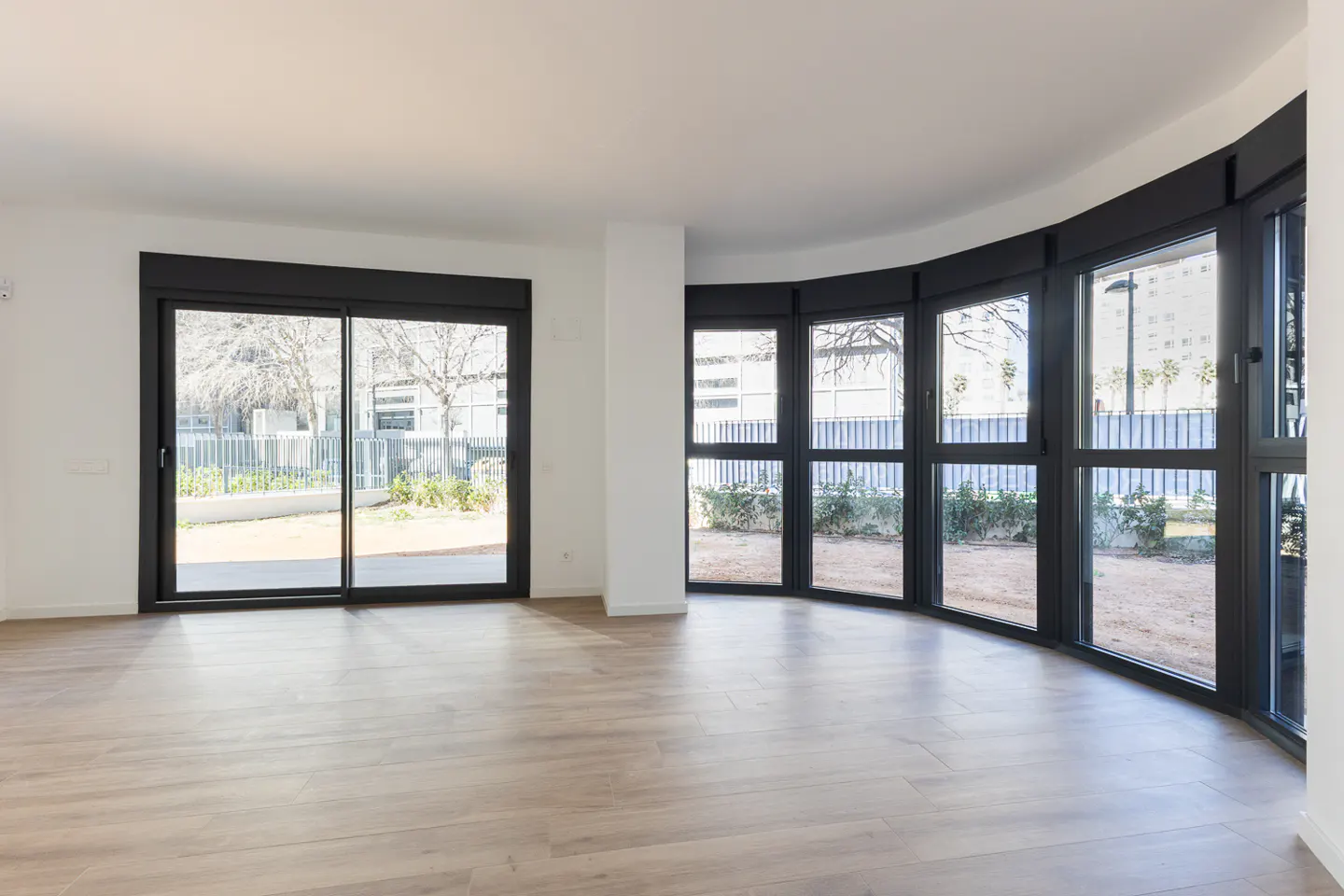 Bright, empty room with light wood floors, white walls, and black-framed windows overlooking a yard with trees and a fence.