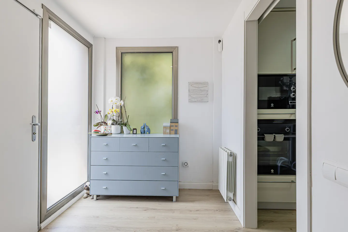 Hallway with a blue dresser, orchids, and a frosted window. A doorway reveals a kitchen with black appliances.
