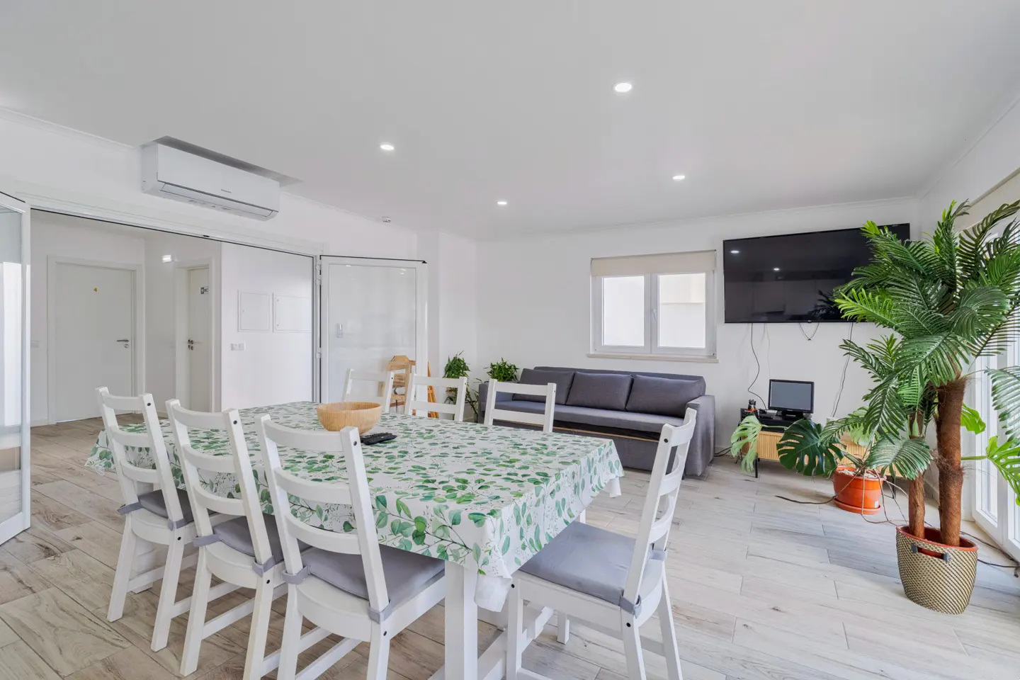 Bright, modern living room with a dining table covered in a green leaf patterned cloth, white chairs, and a gray sofa. A large TV and potted plants are visible.