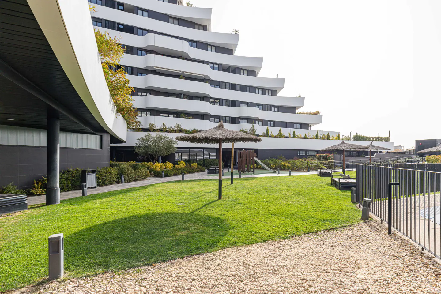 Modern white apartment building with tiered balconies overlooks a green lawn with straw umbrellas and a playground.
