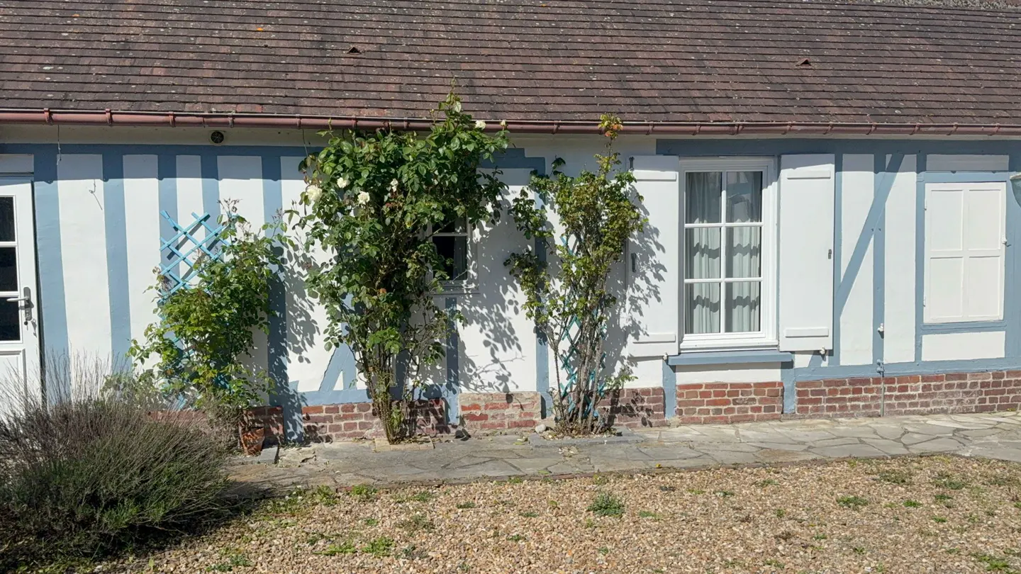 Exterior of a white house with blue trim and a brown roof. Green plants grow near white windows with shutters.
