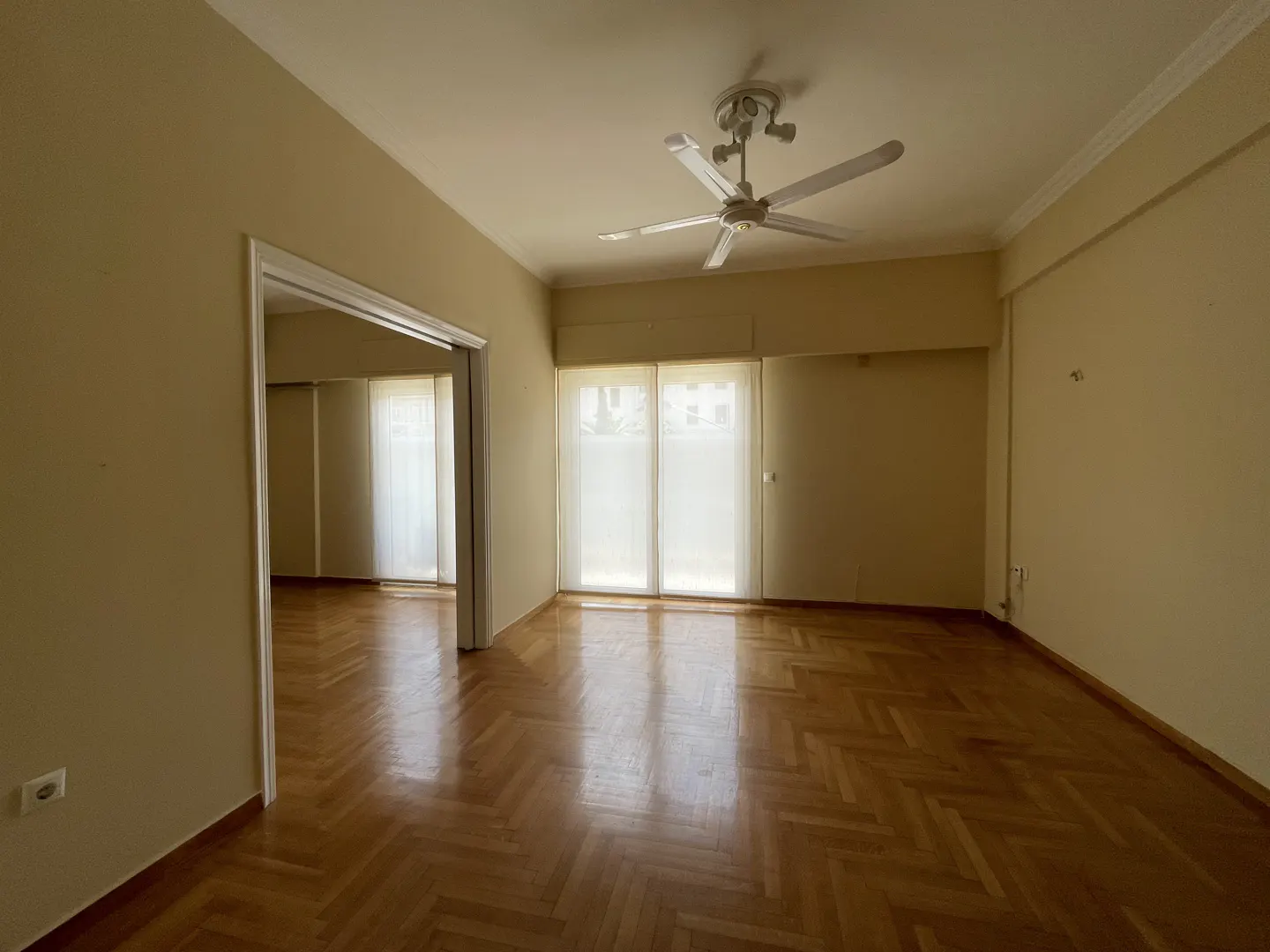 Empty room with herringbone wood floors, cream walls, and a white ceiling fan. Curtains cover the windows.