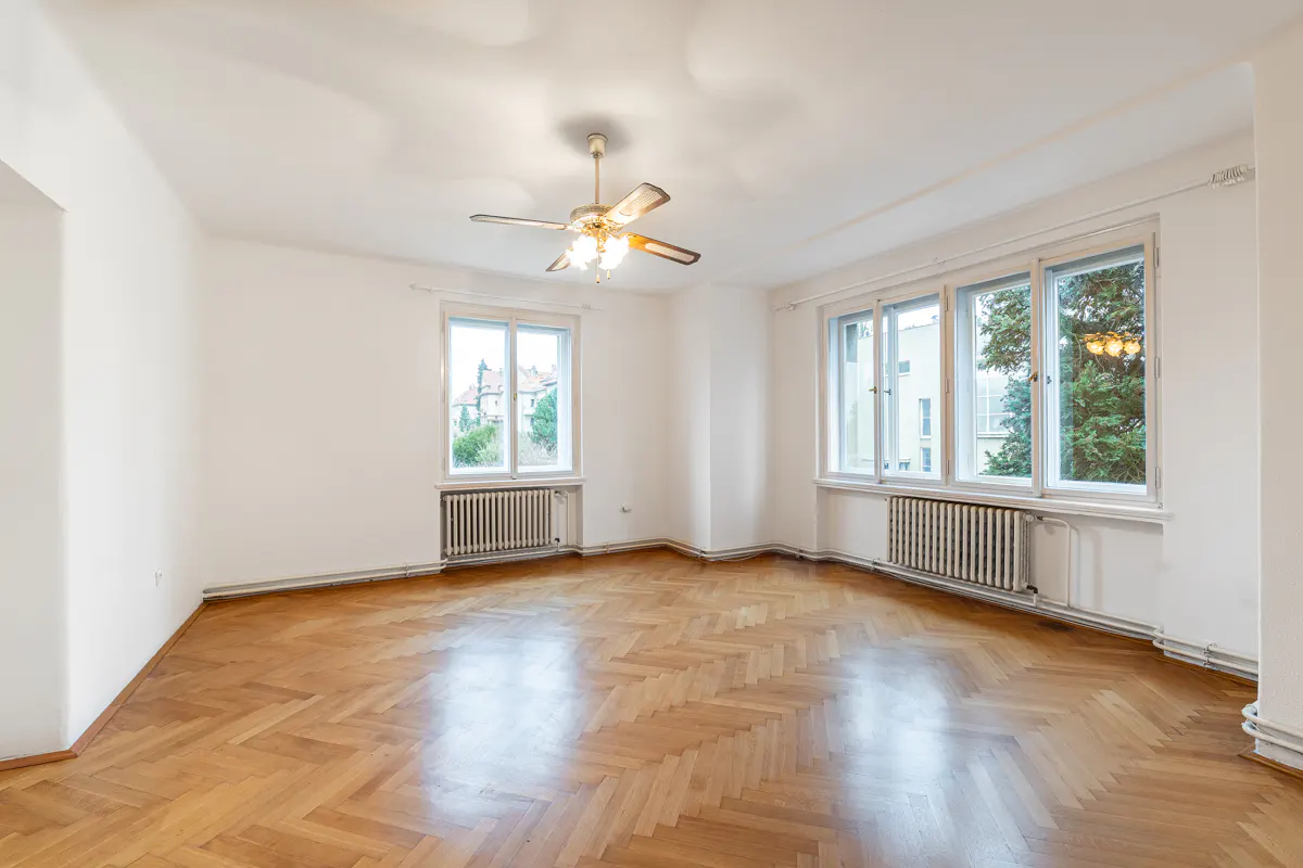 Empty room with white walls, herringbone wood floors, a ceiling fan, and windows with radiators underneath.