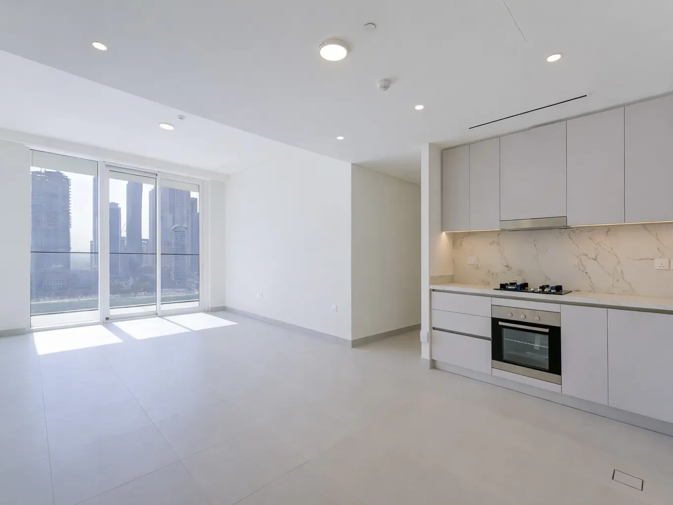 Bright, modern apartment with white walls, tile floor, and a kitchen with gray cabinets. A large window shows a city skyline.