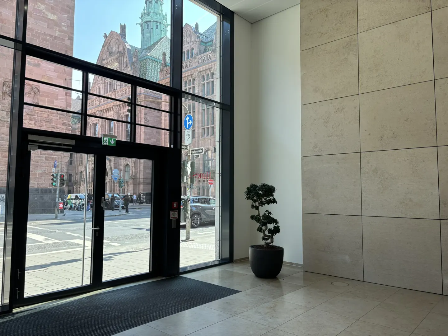 Interior view of a building with a black framed glass door and windows. Outside is a street with a large brick building. A small tree sits in a pot near a tiled wall.
