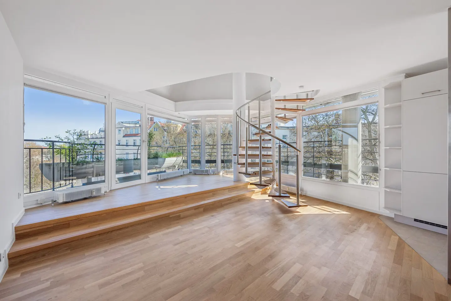 Bright, empty room with wood floors, spiral staircase, and large windows overlooking a balcony and city.