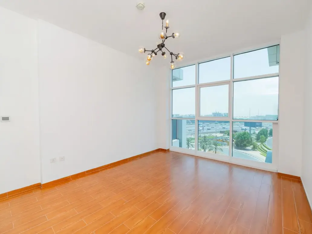 Empty room with wood floors, white walls, and a modern chandelier. Large window shows a city view.