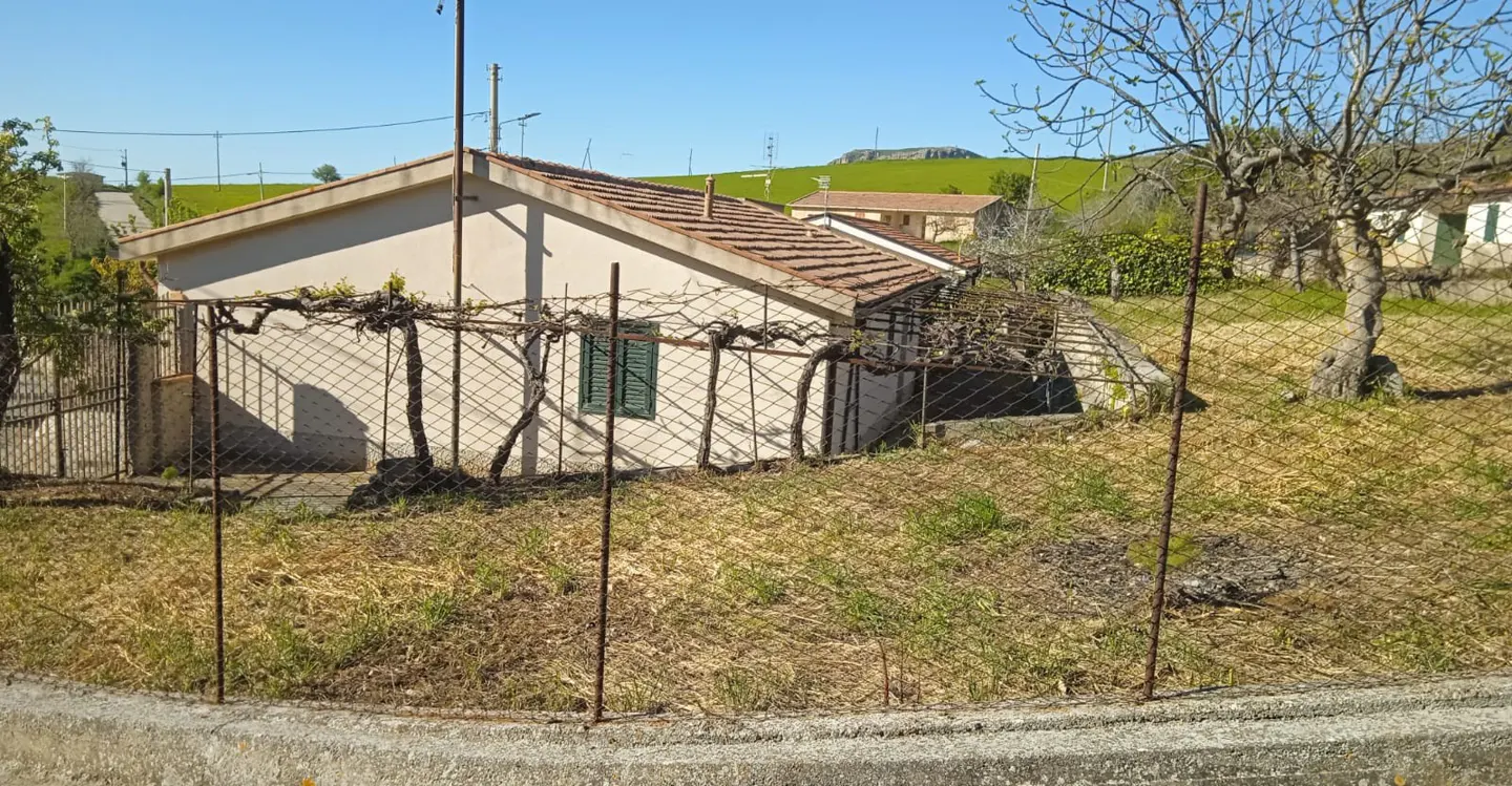 A small, one-story house with a brown tile roof and beige walls, surrounded by a wire fence and dry grass. A vine grows along the side of the house.