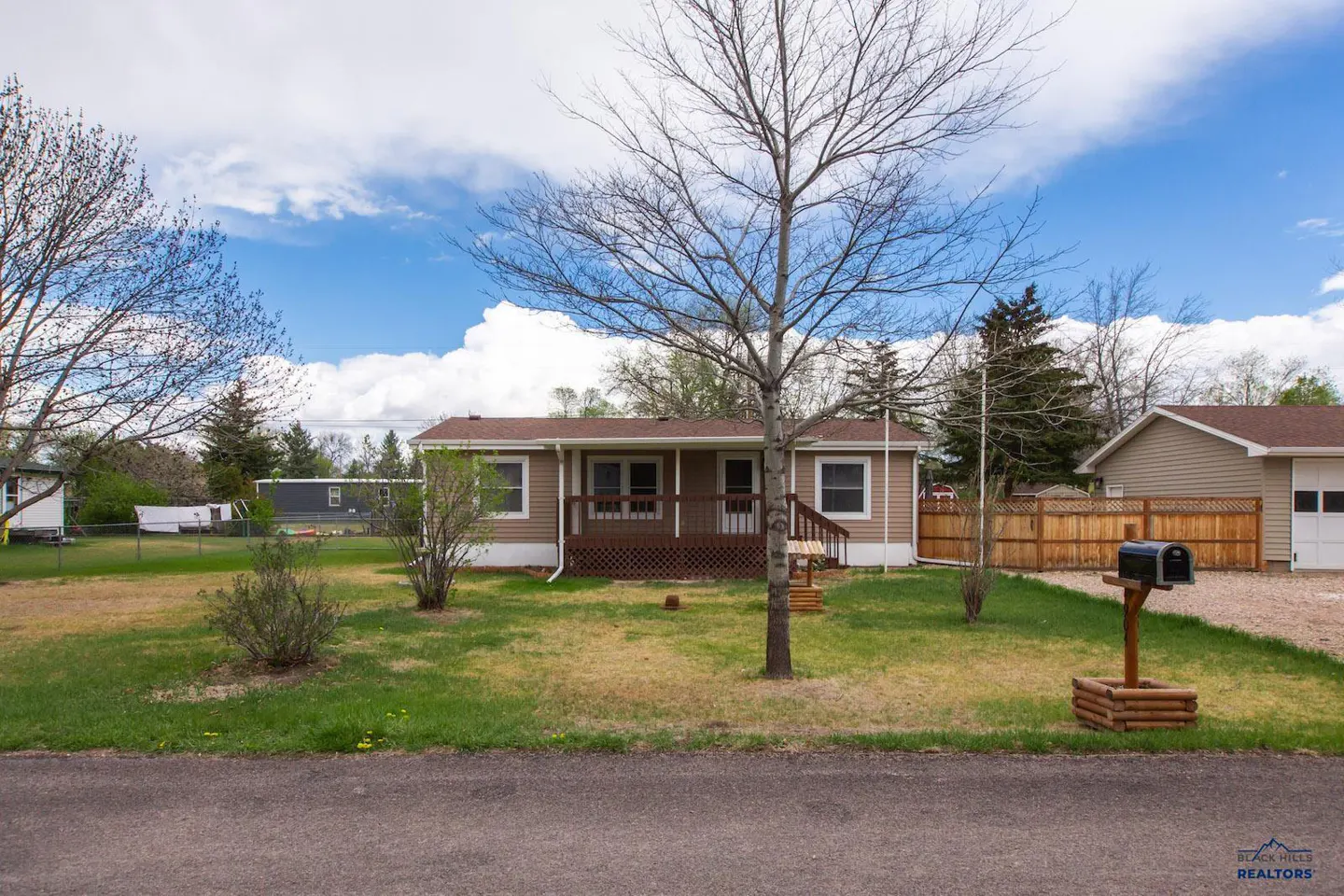 Tan single-story house with a brown roof, porch, and a leafless tree in the front yard under a cloudy sky.