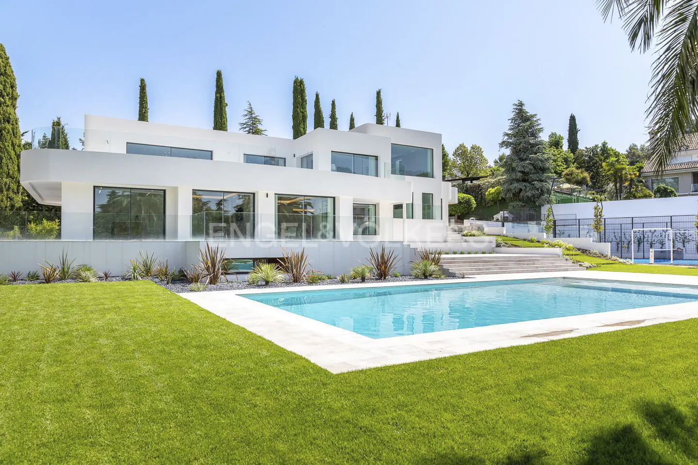 Modern white luxury villa with large windows, a turquoise swimming pool, and a manicured lawn under a clear blue sky.