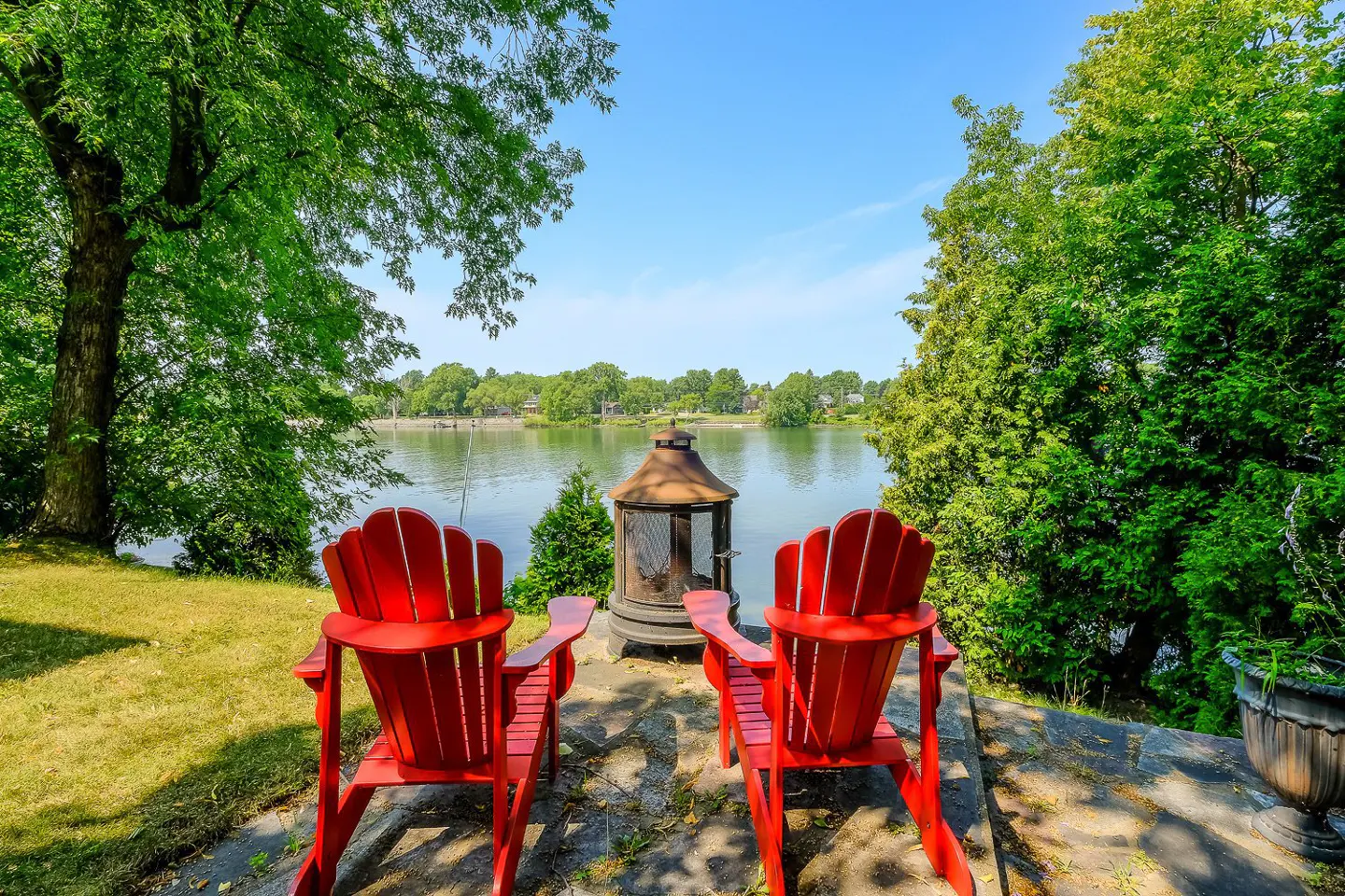 Two red Adirondack chairs face a lake with a fire pit between them, under a blue sky.