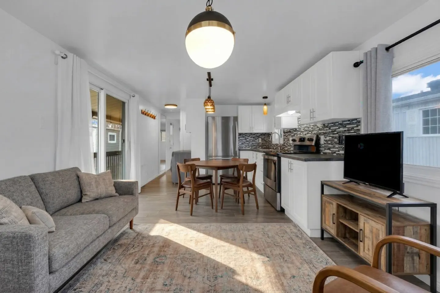 Bright, open-concept living space with a gray sofa, wood dining set, and white kitchen cabinets. A patterned rug adds warmth to the room.
