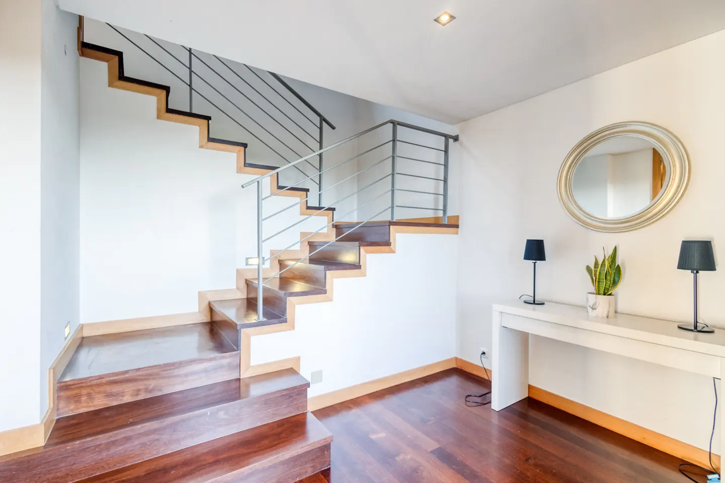 Interior view of a home featuring a wooden staircase with metal railings, a white console table with lamps, and a round mirror.