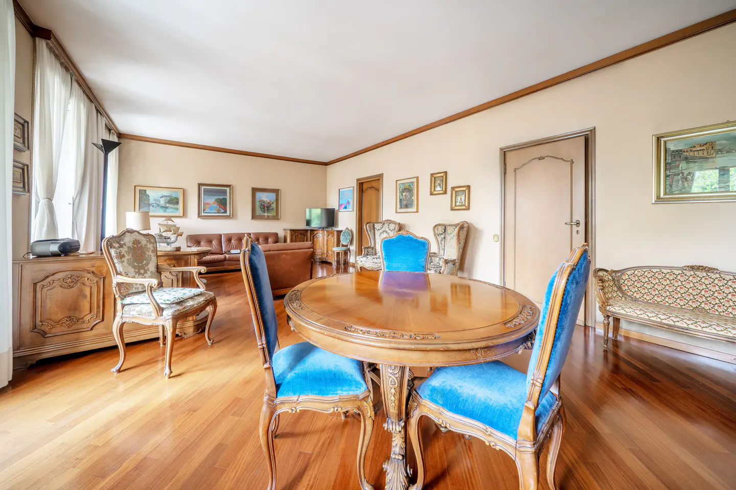 Classic dining room with a round wooden table, blue velvet chairs, hardwood floors, and traditional wall art.