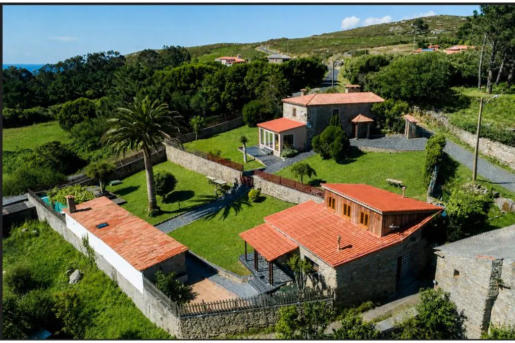 Aerial view of a stone house with a red tile roof, surrounded by green grass, trees, and a stone wall.