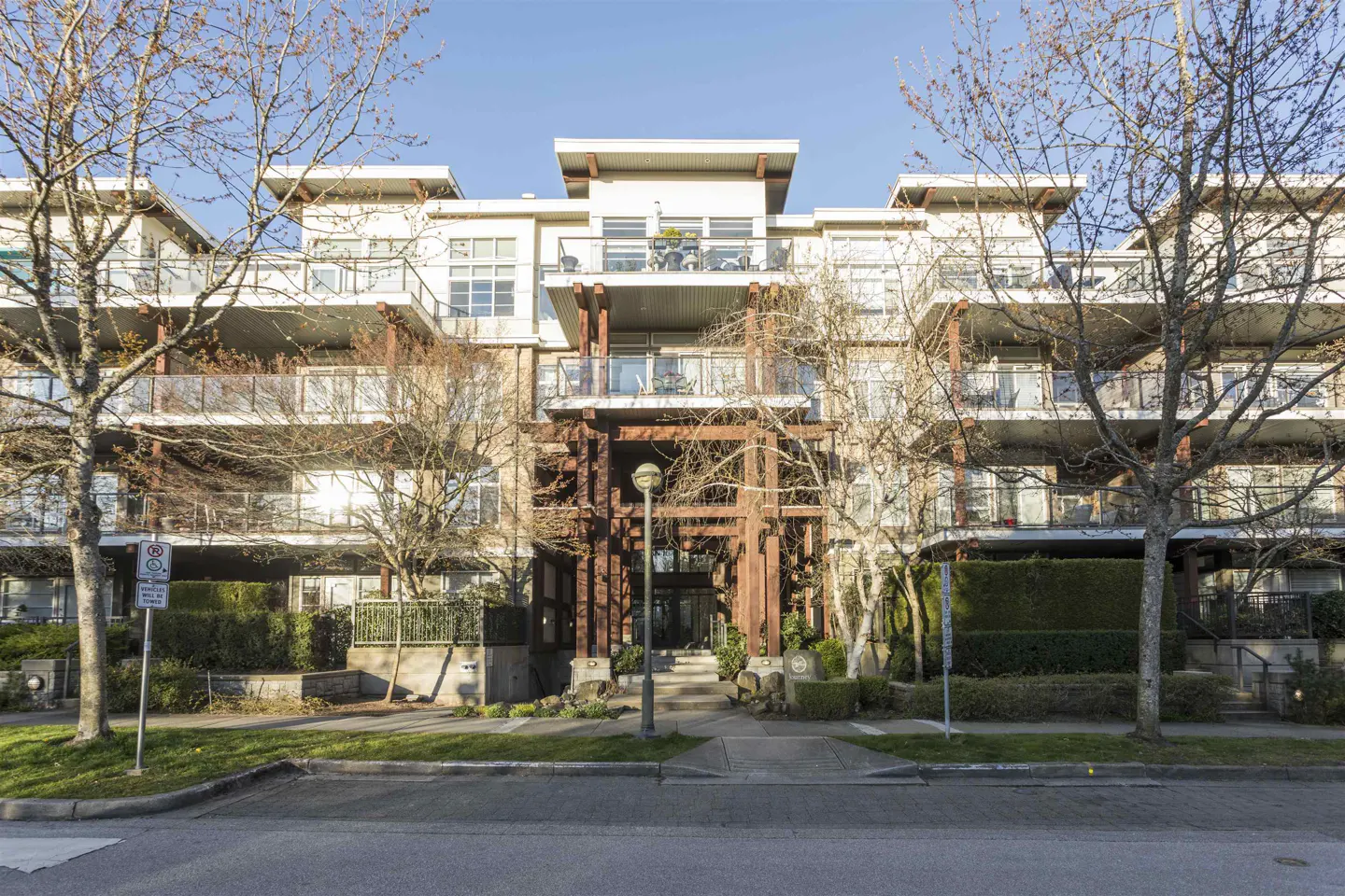 Exterior of a modern, multi-story condo building with balconies, a wood-framed entrance, and trees in the foreground.