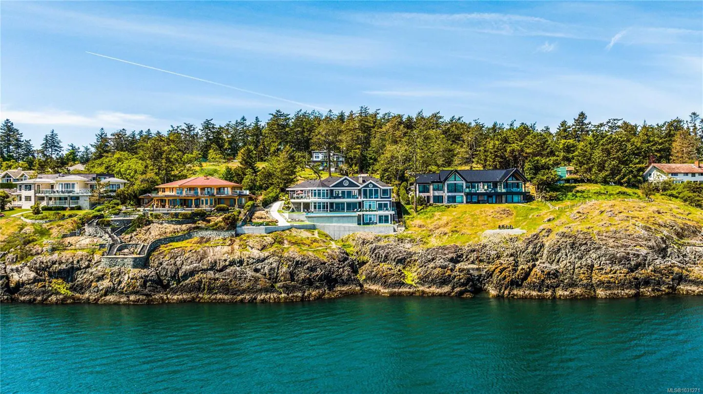 Waterfront homes sit atop rocky cliffs with green trees under a blue sky.