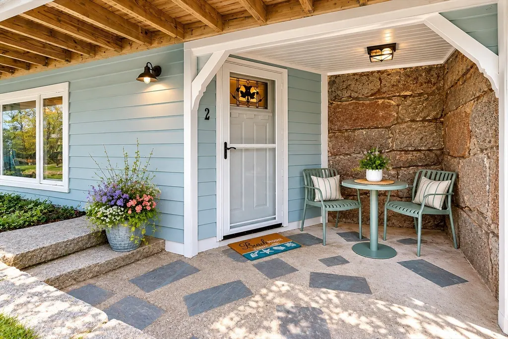 Exterior view of a light blue house with a stone porch, two green chairs, and a small round table. A flower pot sits near the steps.