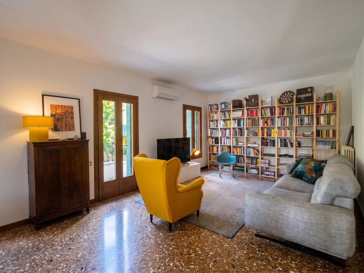 A living room with a yellow armchair, a gray sofa, and a large bookshelf filled with books. The room has a terrazzo floor and a door leading to a balcony.