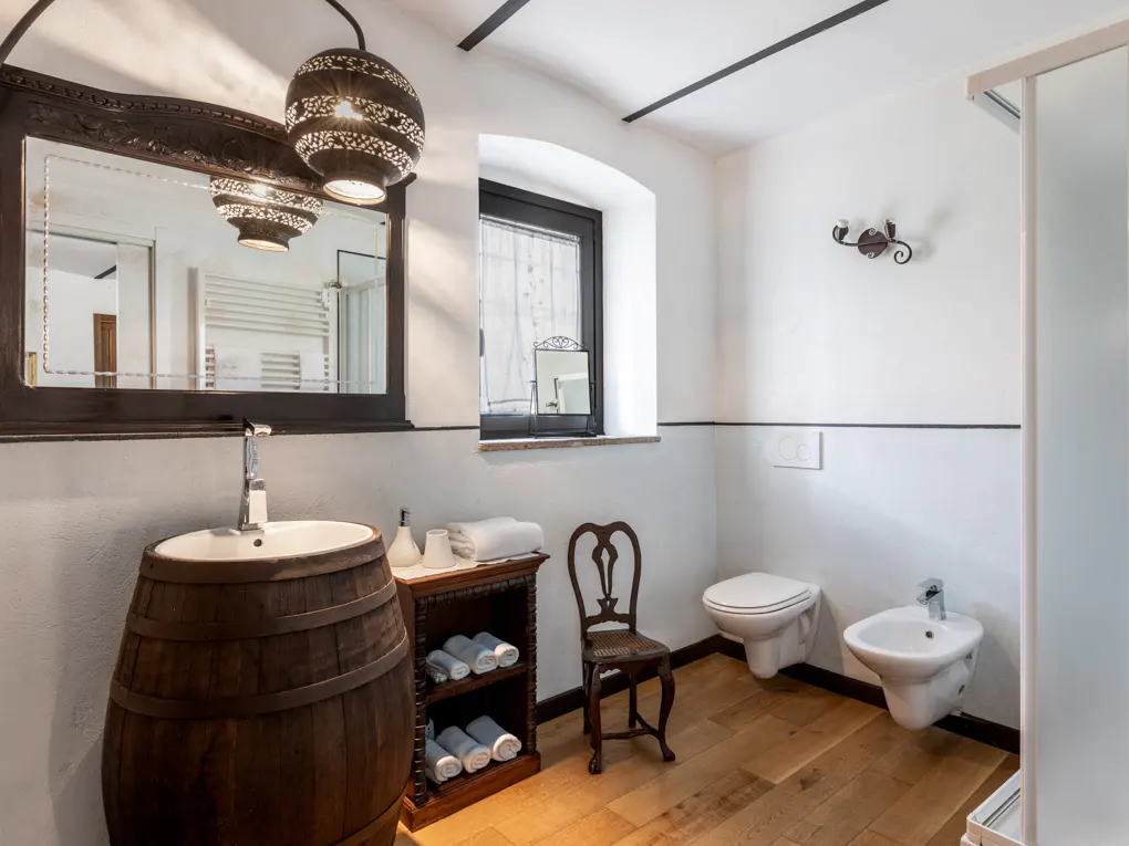 Bathroom with a barrel sink, dark wood mirror, and a decorative light fixture. Toilet and bidet are on the right. White walls and wood floors.