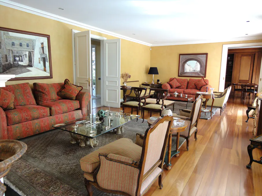 Living room with yellow walls, hardwood floors, and red patterned sofas. A glass coffee table sits on a patterned rug. Artwork hangs on the walls.