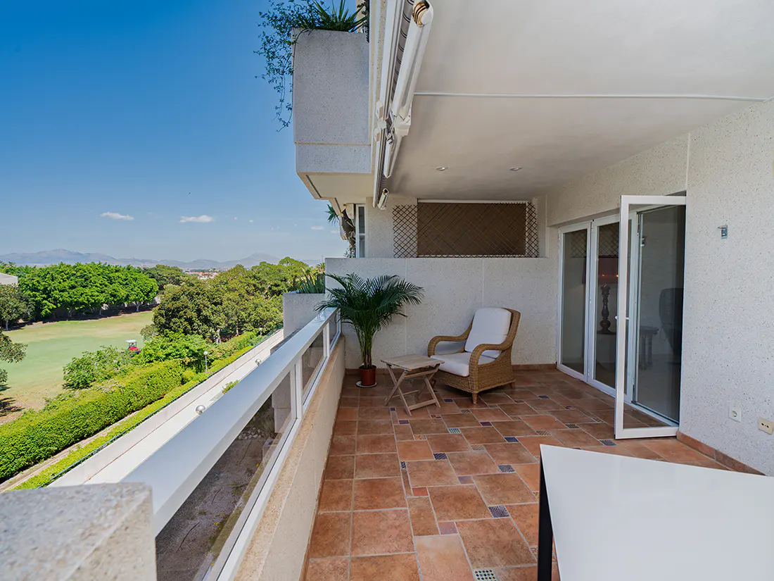 Balcony with brown tile floor, wicker chair, and small table. A potted palm sits near the railing, overlooking green trees and a blue sky.