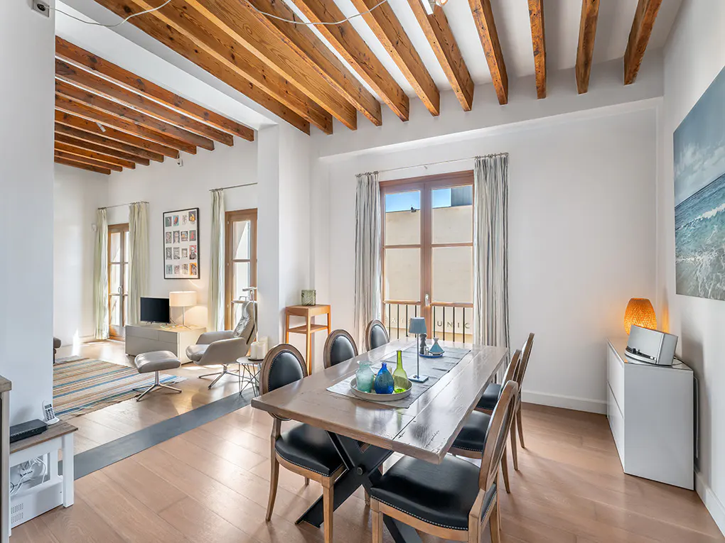 Bright, open-plan dining and living room with wooden beams, table, chairs, and a beach painting.