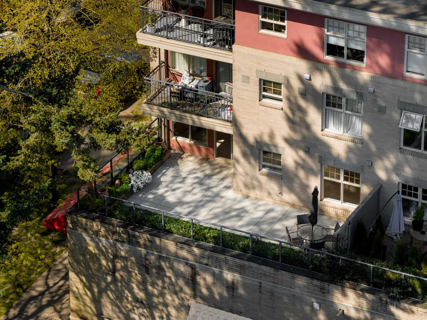 Aerial view of a multi-story apartment building with balconies and a patio. The building is brick with a red accent. Trees surround the building.
