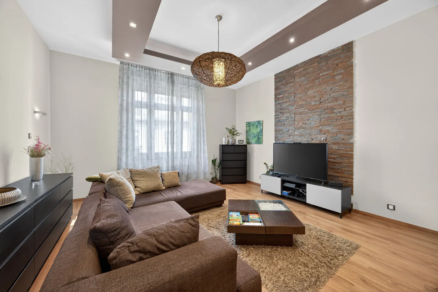Modern living room with a brown sectional sofa, stone accent wall, flat-screen TV, and a unique woven pendant light.