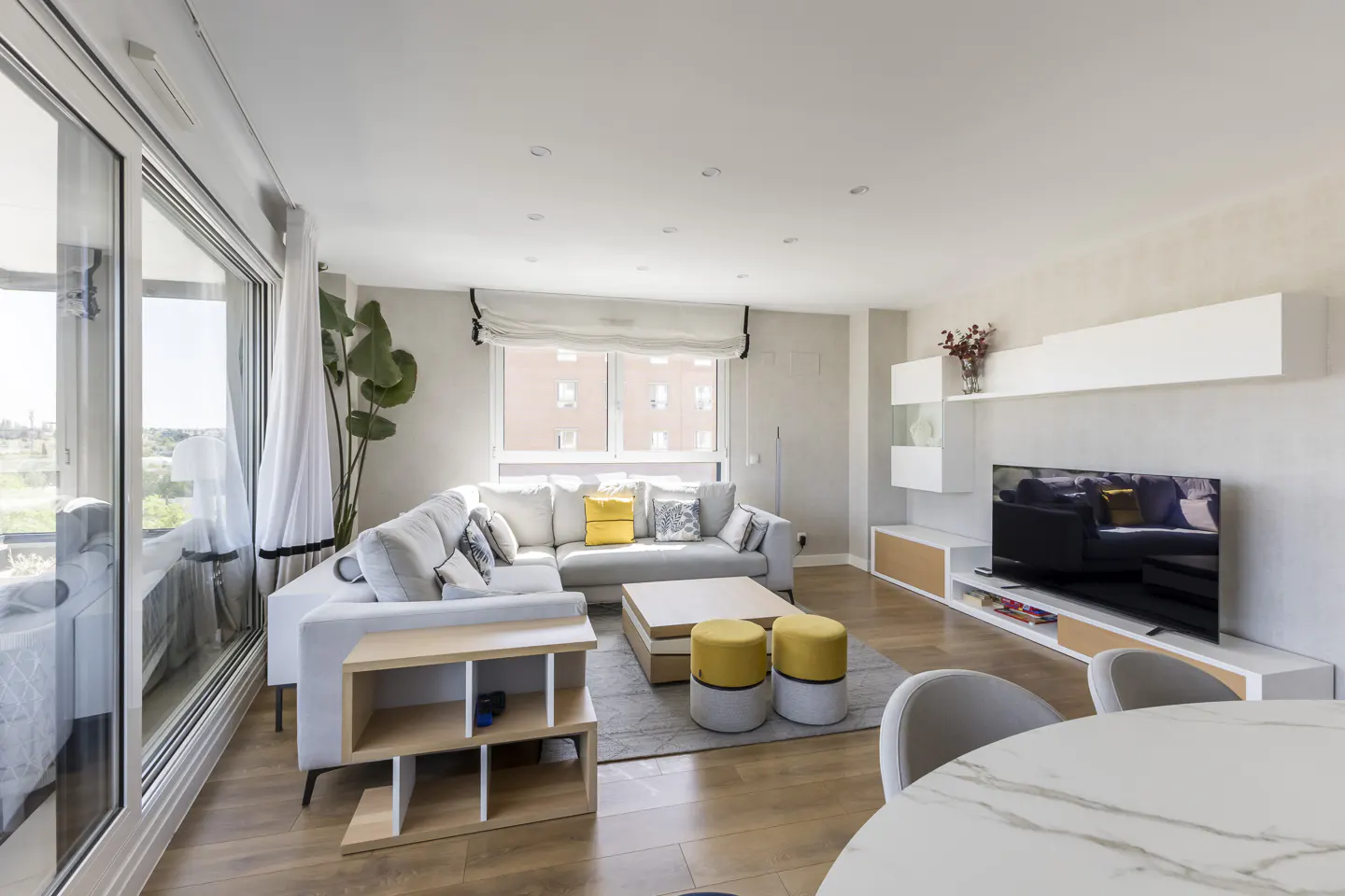 Bright living room with a white sectional sofa, yellow accents, and a large window. A TV and modern shelving unit are on the far wall.