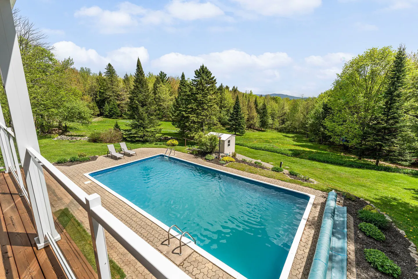 Aerial view of a rectangular in-ground pool with blue water, surrounded by a brick patio, green lawn, and trees. Two lounge chairs are near a shed.