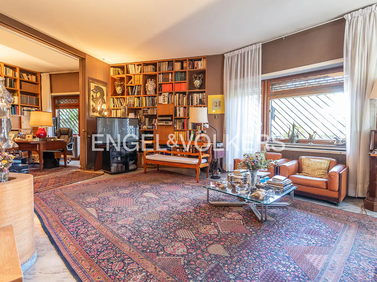 Living room with a large patterned rug, bookshelves, brown walls, and a glass coffee table. Two leather chairs sit near a window with white curtains.