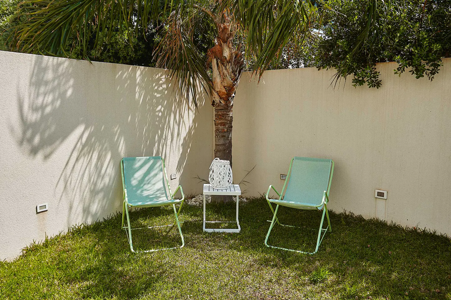 Two light green folding chairs and a small white side table with a lantern sit on a lawn against a beige corner wall.