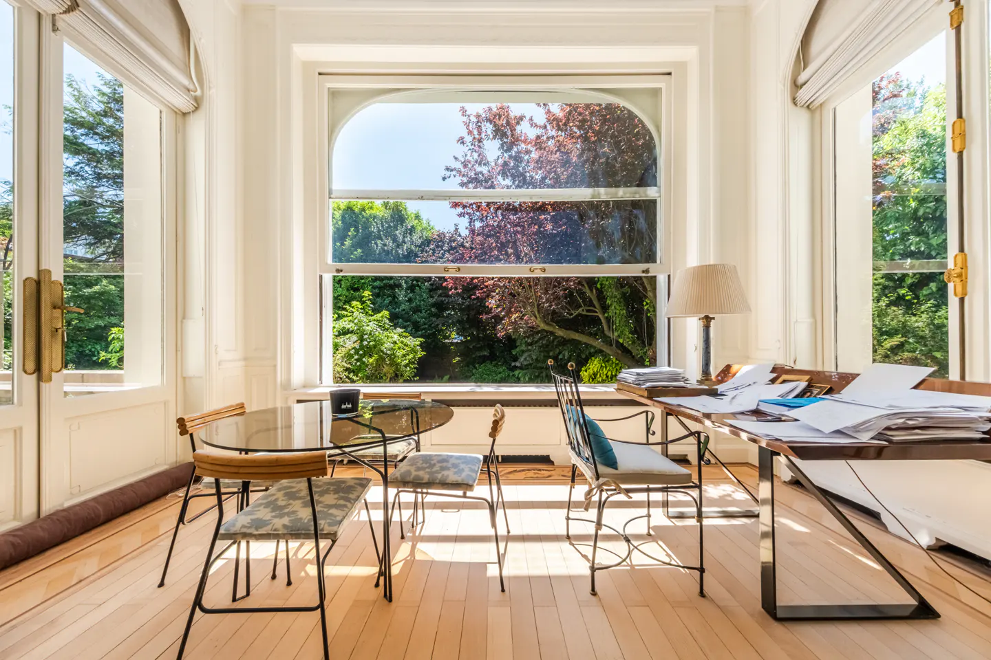 Bright sunroom with a glass table, chairs, and a desk piled with papers. Large windows overlook a lush green garden.