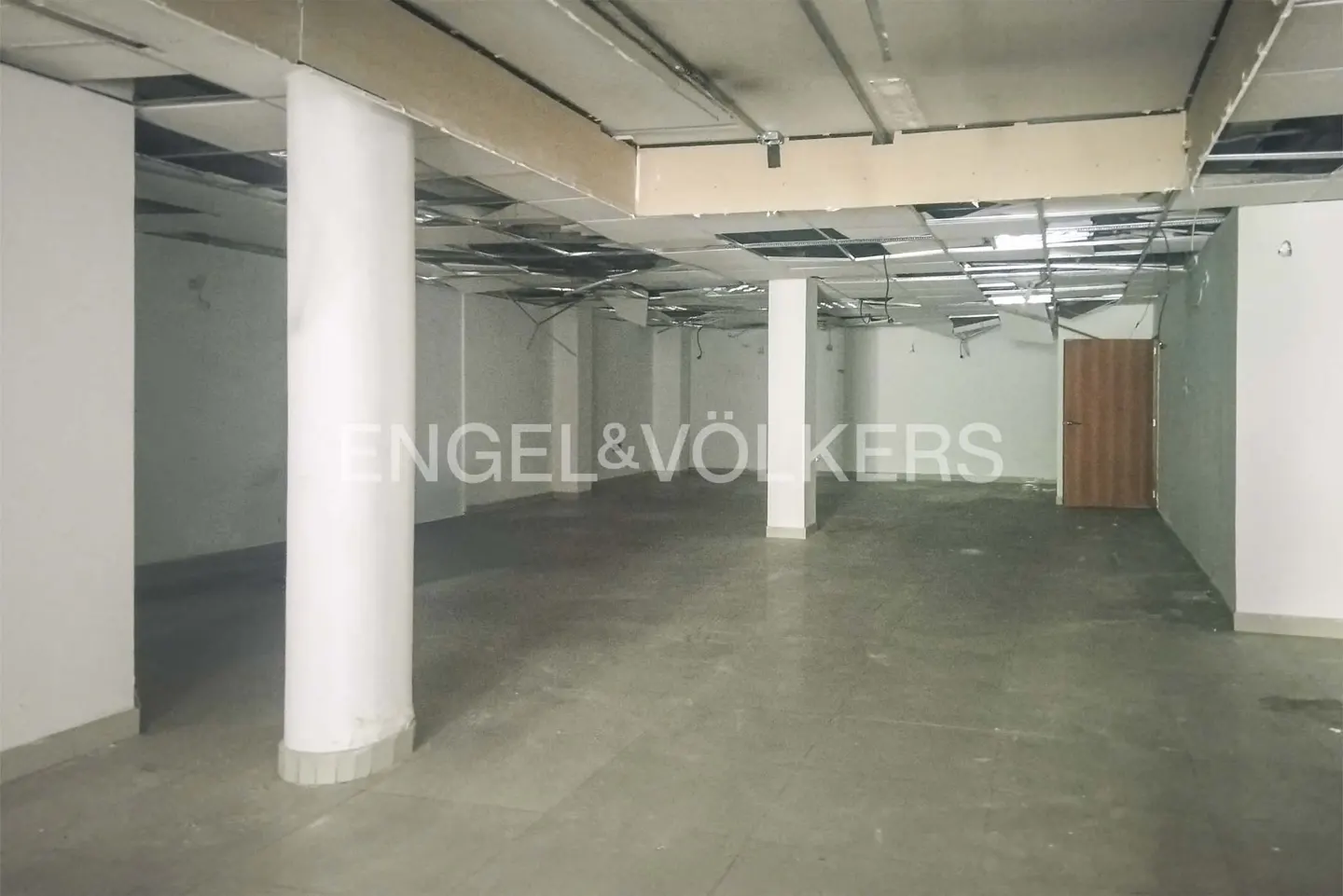 An empty commercial space with white walls, concrete floors, and exposed ceiling with support columns. A brown door is visible in the background.